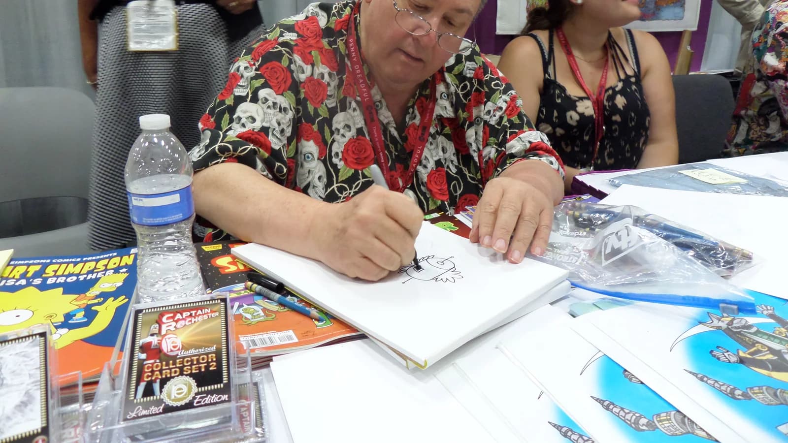 An artist drawing in a sketchbook at a convention table stacked with comics, other people seated alongside.
