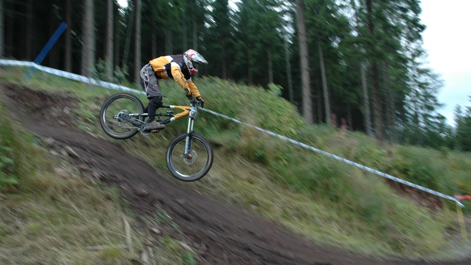 A rider in a full-face helmet and body armour airborne off a steep rutted forest track, both wheels clear of the ground, tape marking the course through the trees.