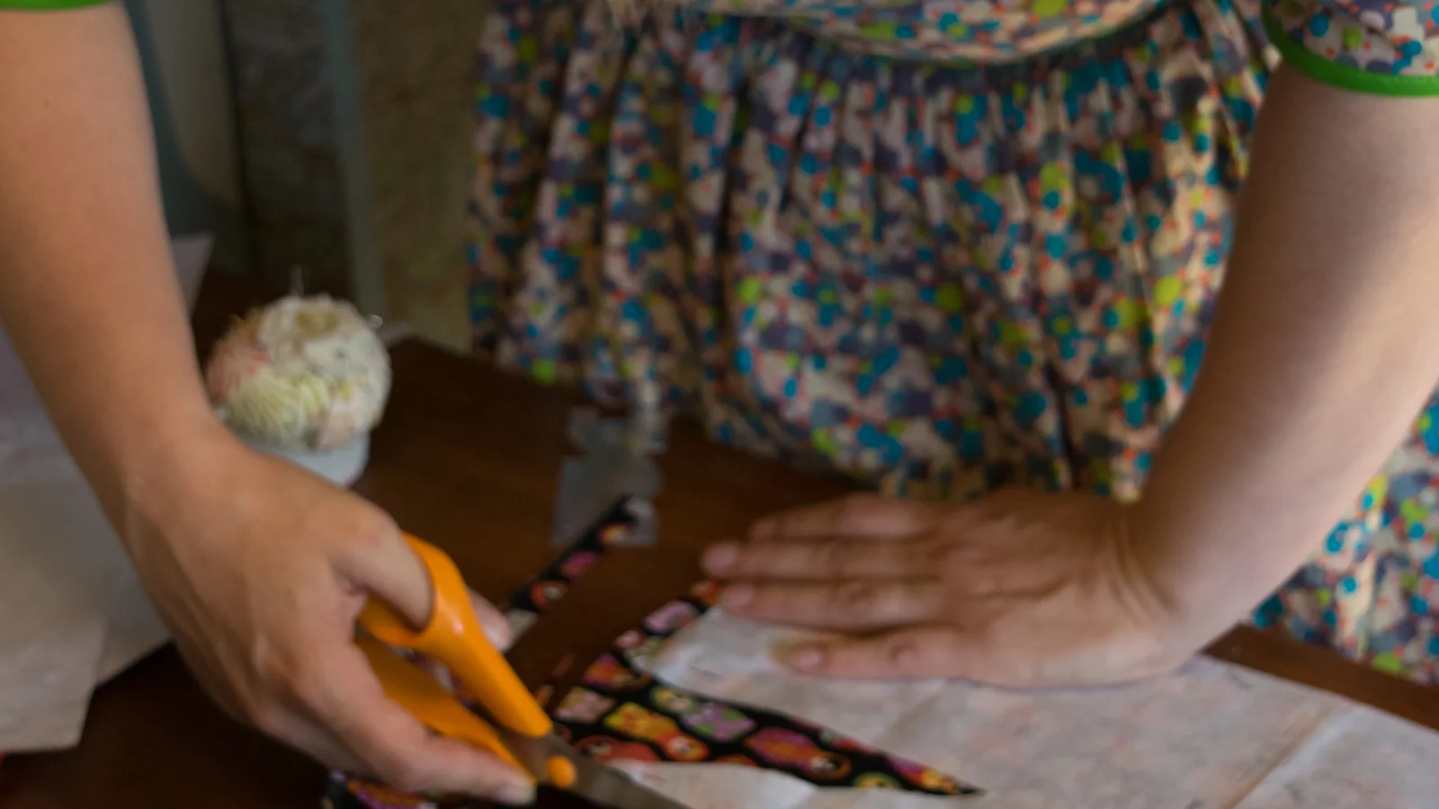 Two hands cutting patterned cotton with orange-handled shears on a table, a pincushion beside them and a printed border running under the blades.