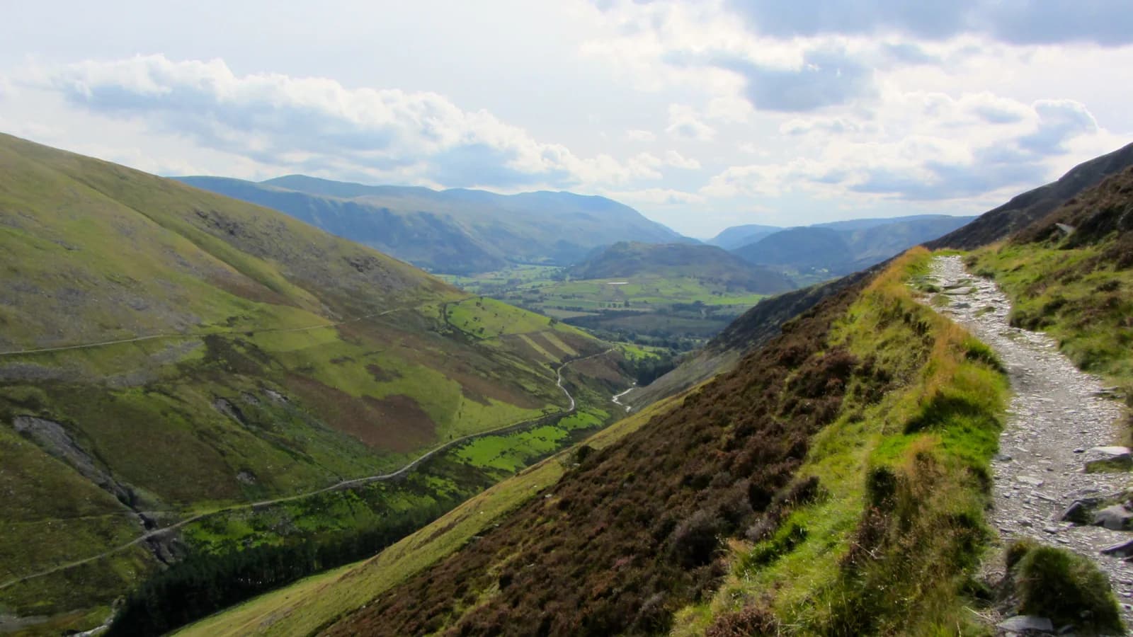 A stony path traversing a steep fellside above a green Lakeland valley, with rough grass and heather on the slope and more fells beyond.