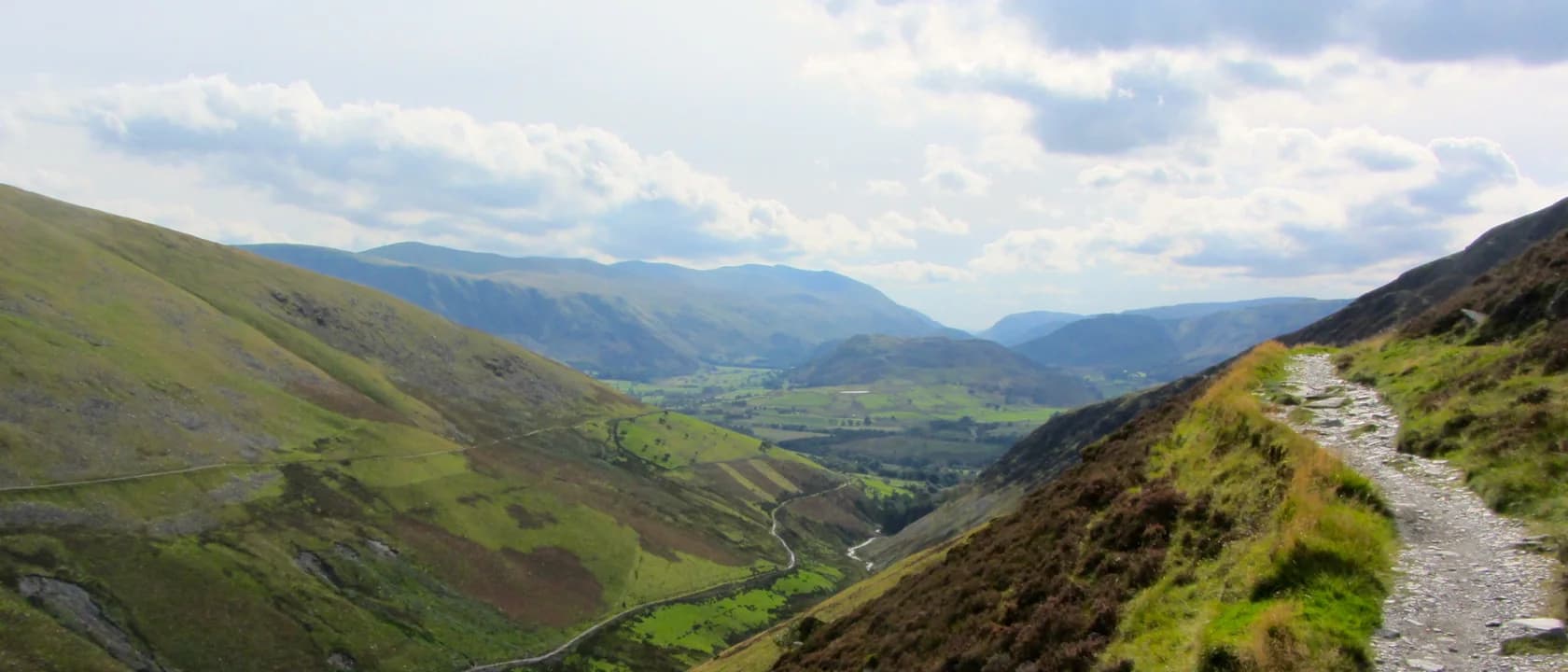 A wide view along a stony path traversing a steep Lakeland fellside, with bracken and heather below it, a green valley far beneath, and further fells receding into haze under a cloudy sky.