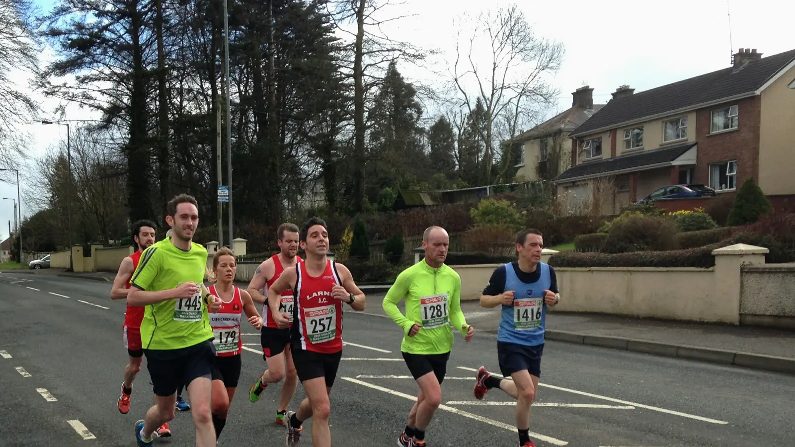 A tight group of eight club runners in numbered vests on an ordinary suburban road, houses and bare trees behind them, nobody watching.