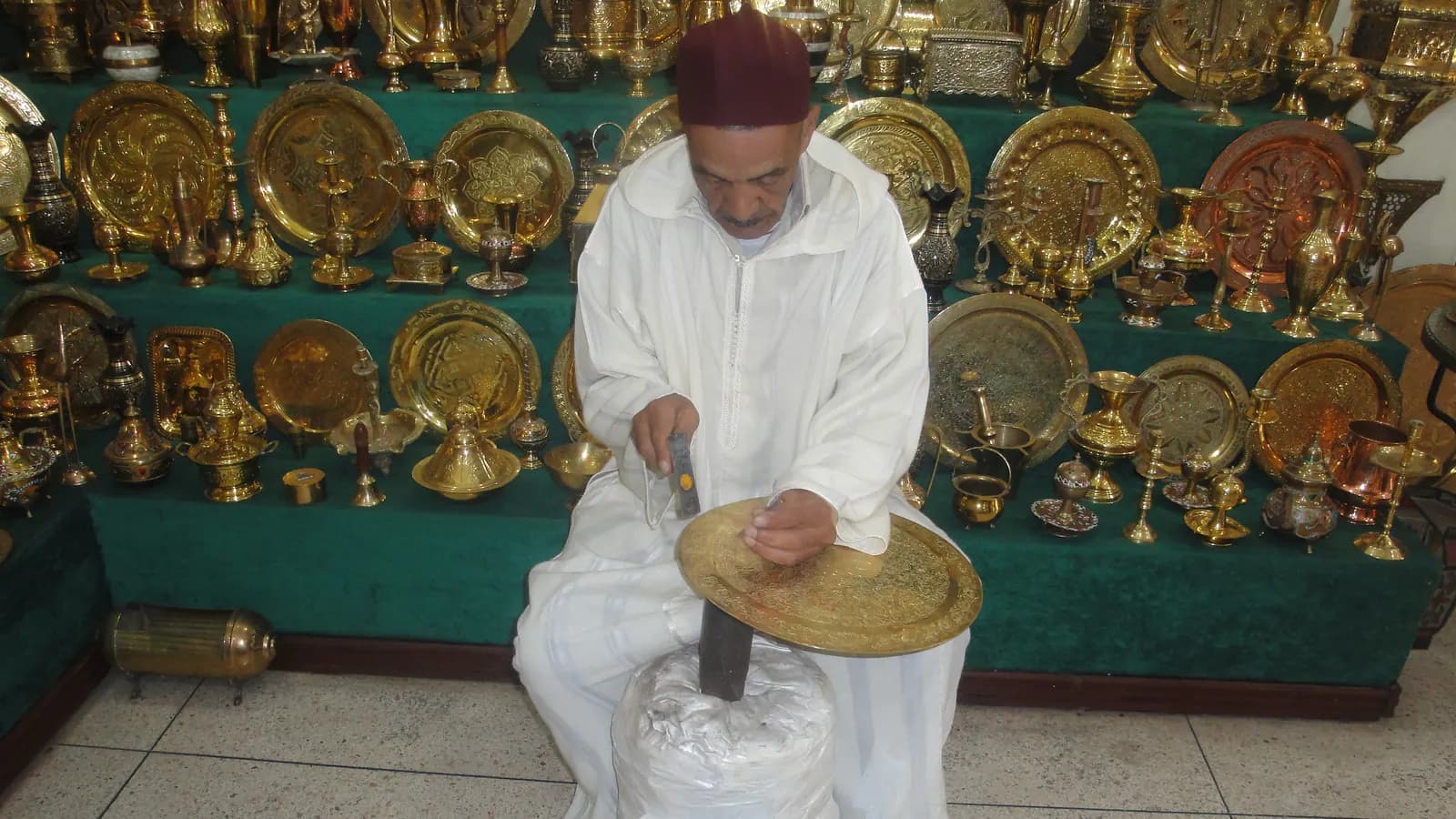 A craftsman seated on the floor chasing a pattern into a large brass tray, with hundreds of finished trays hung on the wall behind him.
