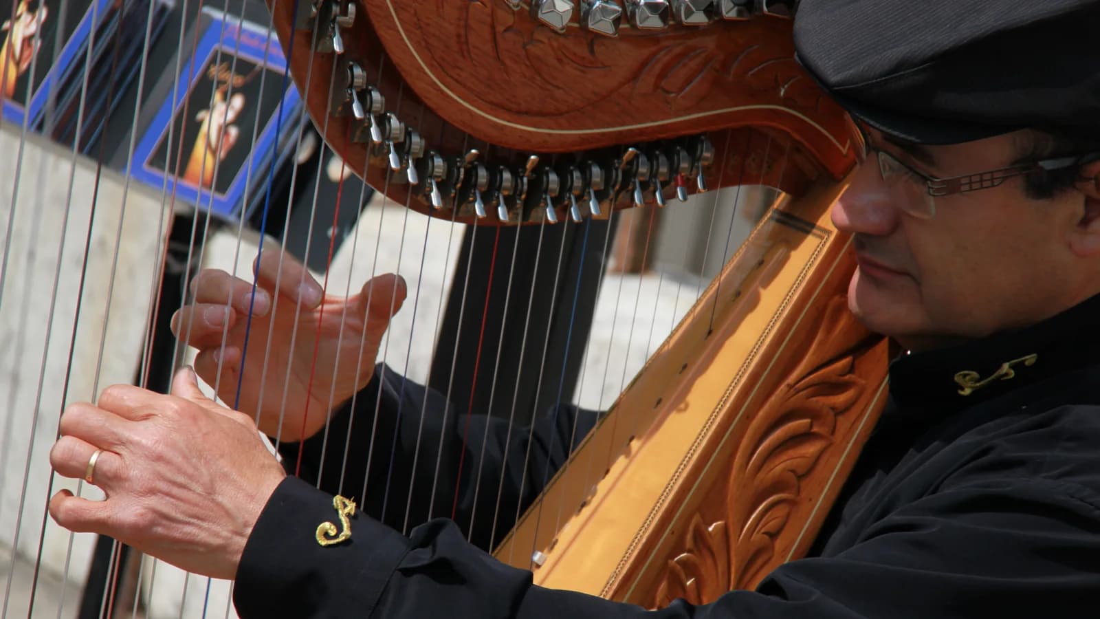 A harpist's hands on the strings of a carved wooden harp, the player in profile behind them wearing a cap.