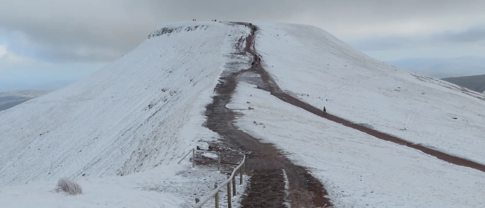 A snow-covered ridge path climbing towards a rounded summit under grey cloud, with a fence line alongside and a walker small on the slope.