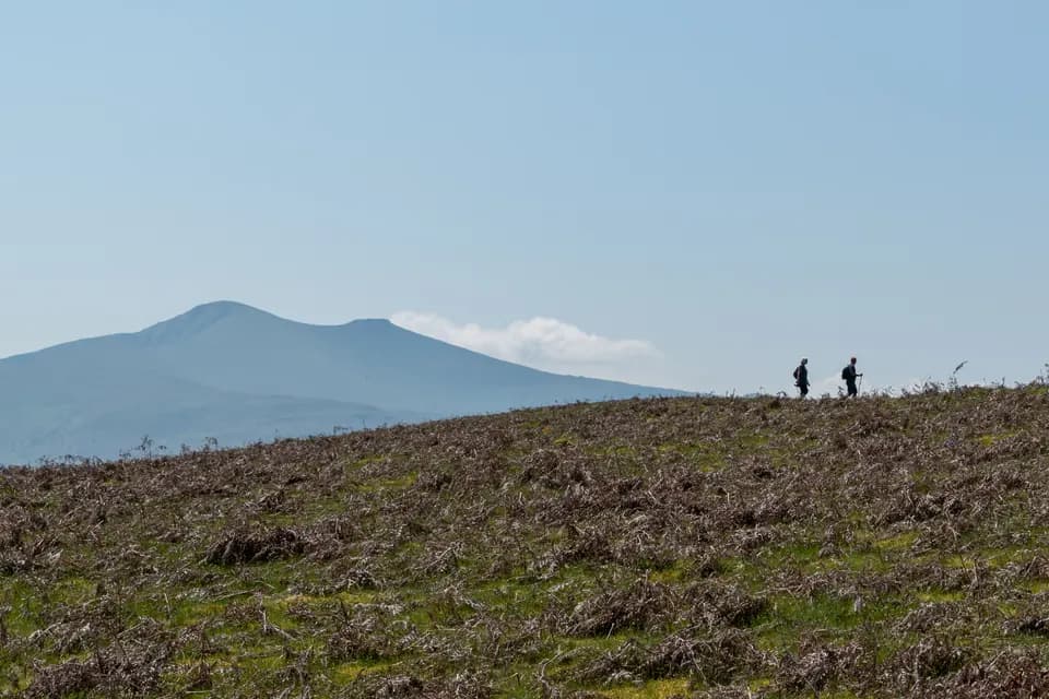 Two walkers on a rough grassy hillside silhouetted against a pale sky, with two flat-topped peaks rising in the distance behind them.