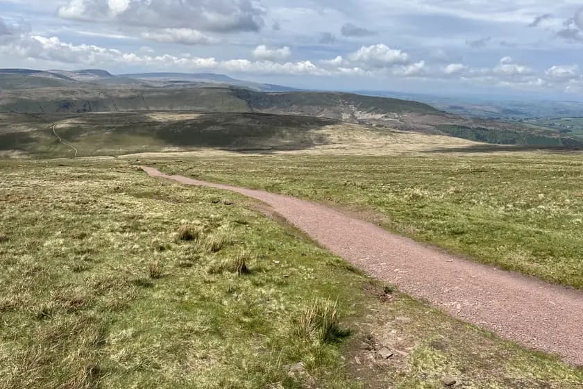 A worn stone path descends across open moorland from high ground, with a wide green valley and distant ridges under a cloudy sky.