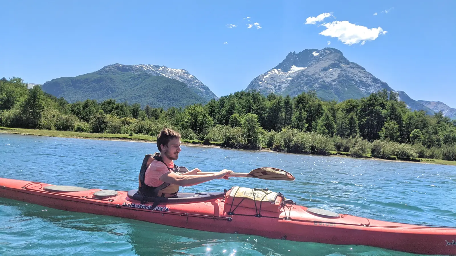 A paddler in a red sea kayak on clear turquoise water, mid-stroke, with wooded banks and a snow-streaked mountain peak behind.