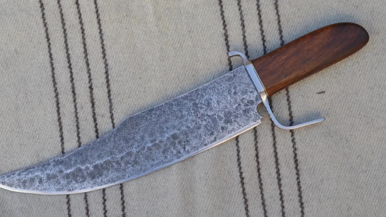 A finished hand-forged bowie knife lying on cloth: a wide blade showing a mottled pattern along its length, a mild steel guard, and a polished hardwood handle.