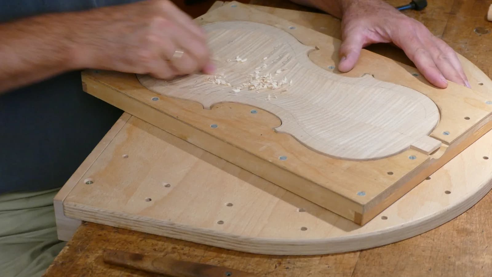 Two hands work a violin-shaped wooden plate clamped to a shaped board on a workbench, with pale shavings scattered around it.