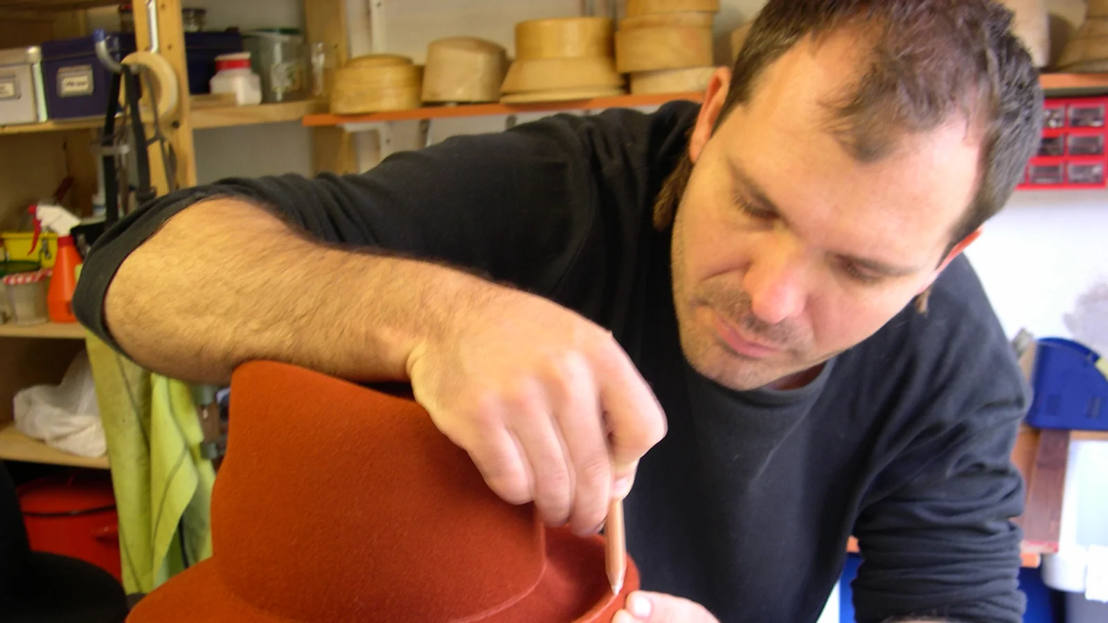 A hat maker in a workshop shaping a red felt hat over a wooden block, with more blocks stacked on shelves behind him.
