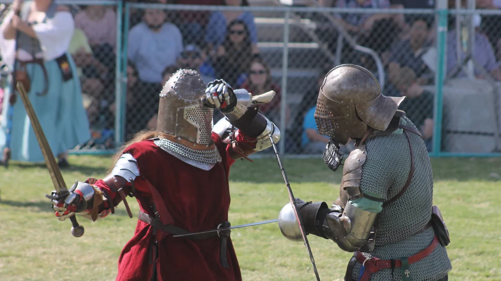 Two live action roleplayers in mail and plate armour fighting with sword and shield on grass, one in red and one in grey, watched by other costumed participants from behind a fence.