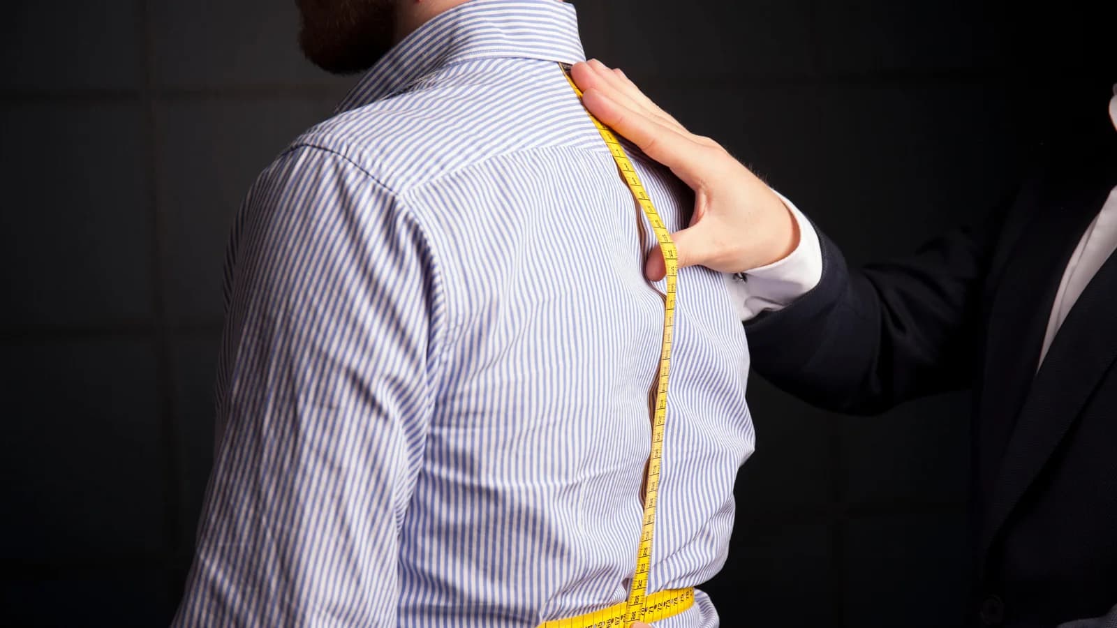 A tailor taking a chest measurement over a striped shirt with a yellow tape, hands positioning the tape at the shoulder.