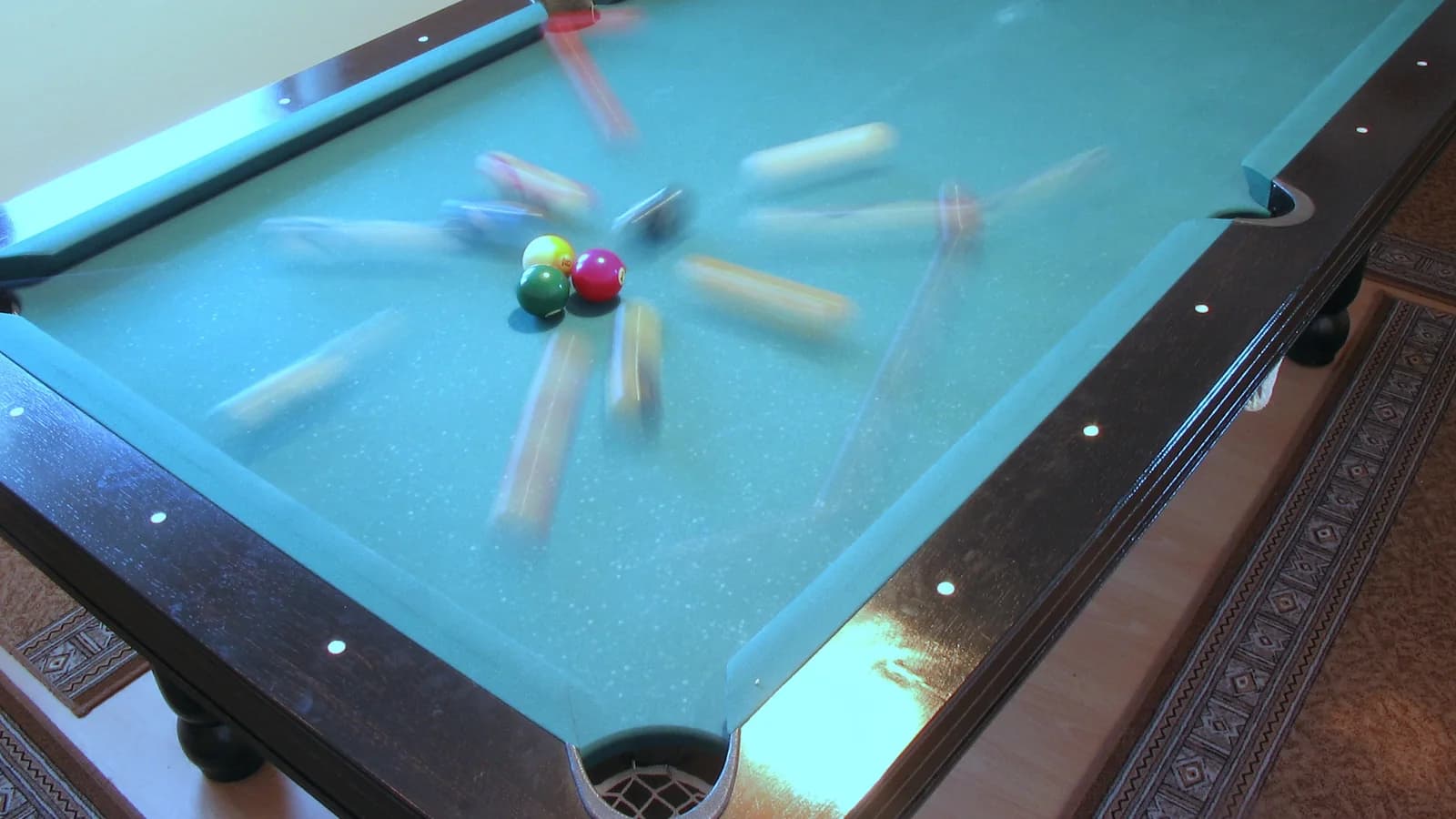 A pool table at the moment of the break, photographed with a long exposure so the scattering balls draw pale streaks across the blue cloth while the few already at rest stay sharp.