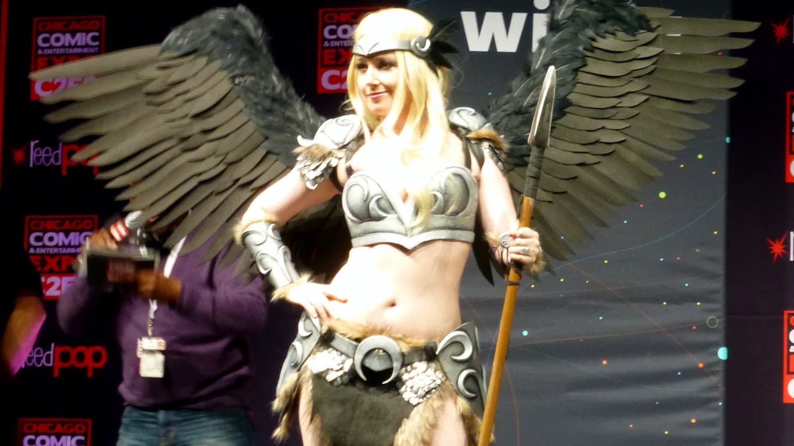 A costumed figure in moulded armour with large feathered wings at a comics convention, photographed against a banner wall.