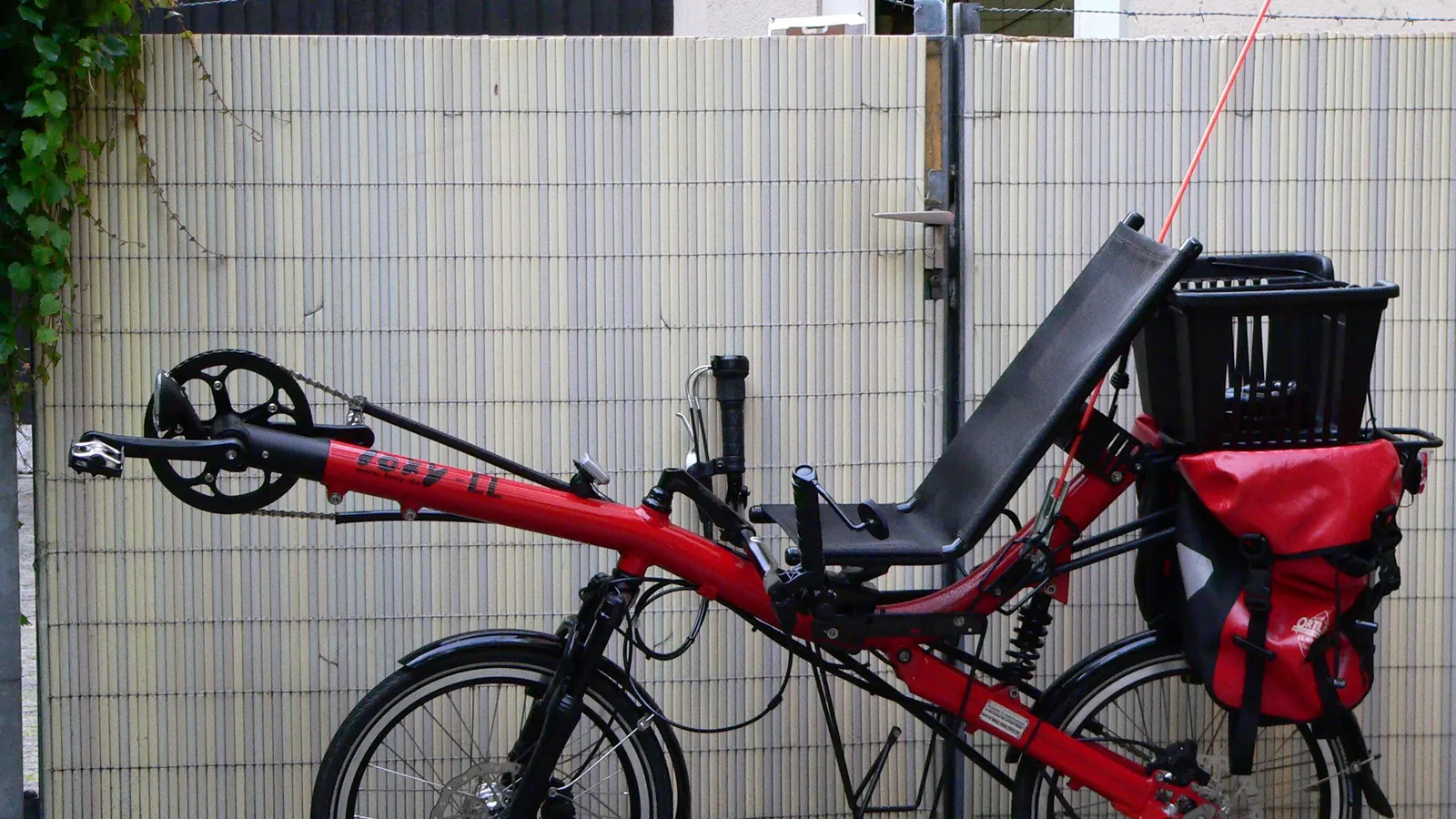 A red short-wheelbase recumbent bicycle seen from the side: a reclined mesh seat, the cranks carried out in front on a long boom, a small front wheel and a larger rear wheel, fitted with panniers and a rear basket.