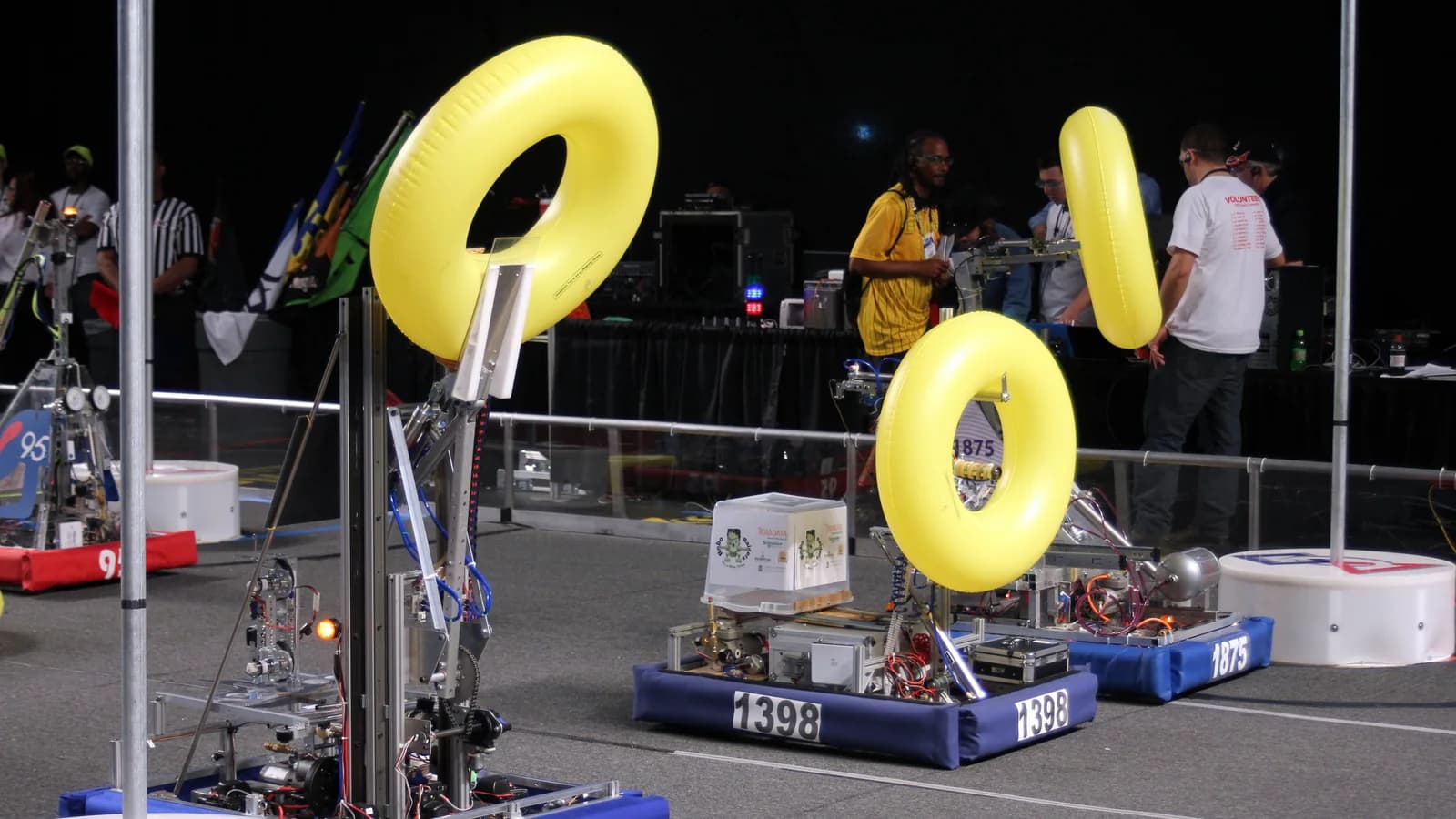Two small competition robots holding large yellow rings on a match field, with team members watching from behind a barrier.