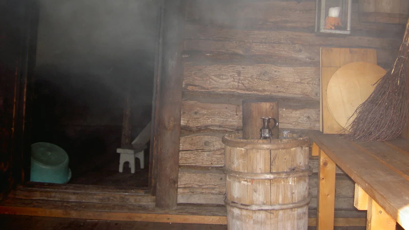 A steamy timber sauna interior with a wooden bucket, a bench and a birch whisk hanging on the wall.
