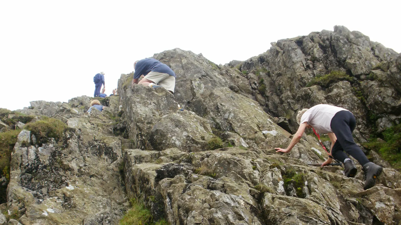 Three people scrambling up a broken rocky ridge, using their hands on the rock as well as their feet, with the crest above them against a pale sky.