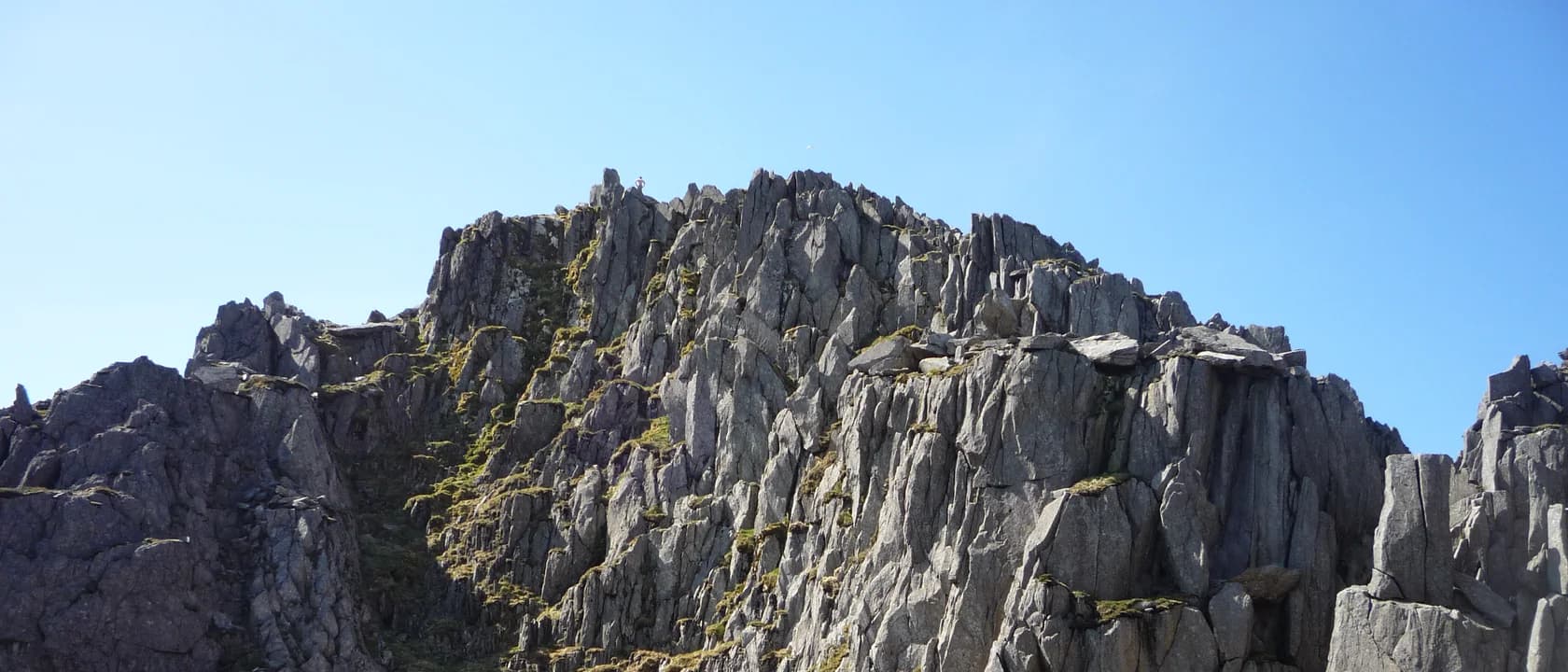 A wide view of a rocky buttress on Tryfan: shattered volcanic rock forming blocks, steps and towers, split by a steep grassy gully, with moss and grass growing in every ledge and crack.