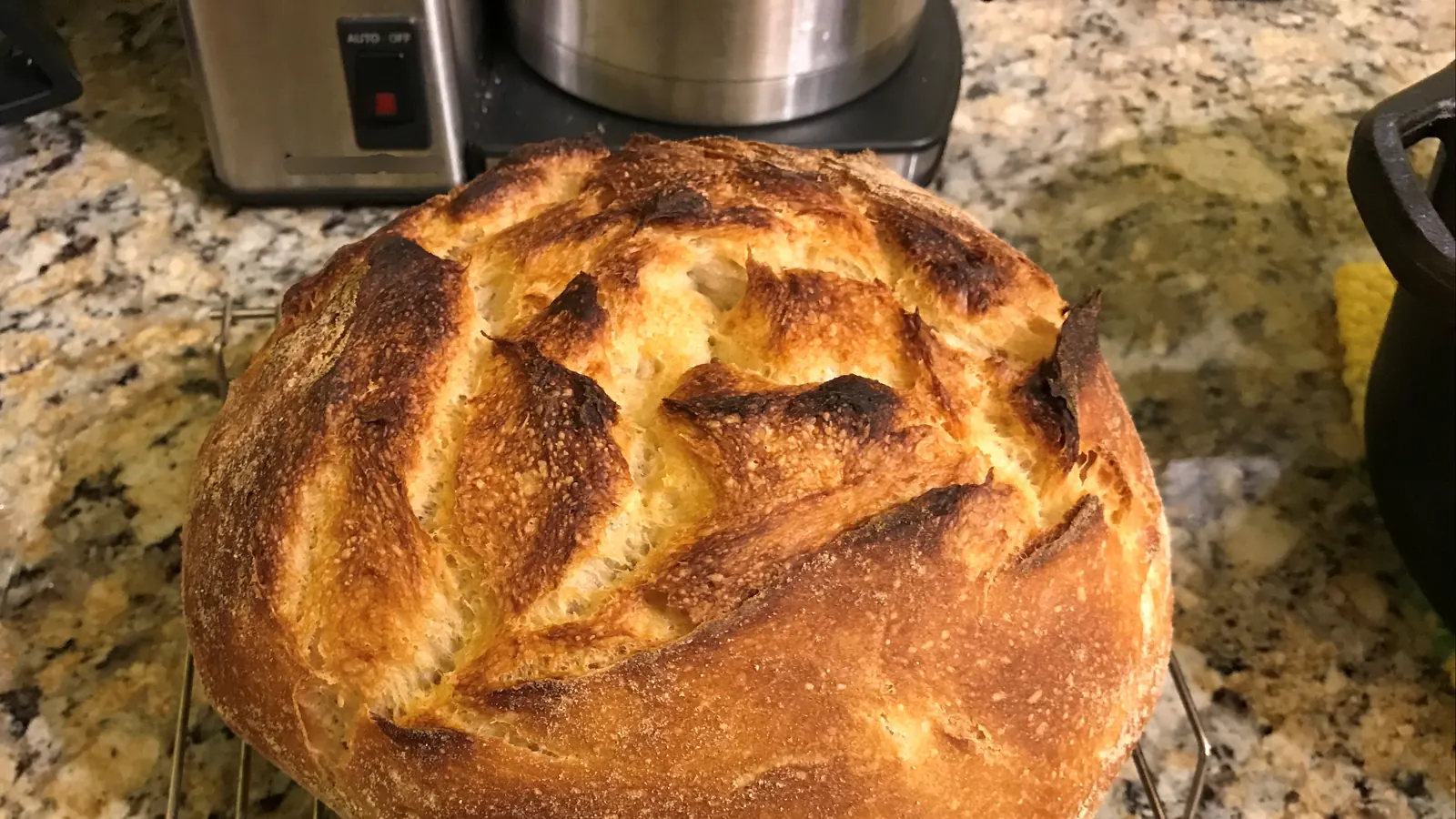 A round sourdough loaf with a deeply browned crust and a spiral scoring pattern, cooling on a stone worktop.