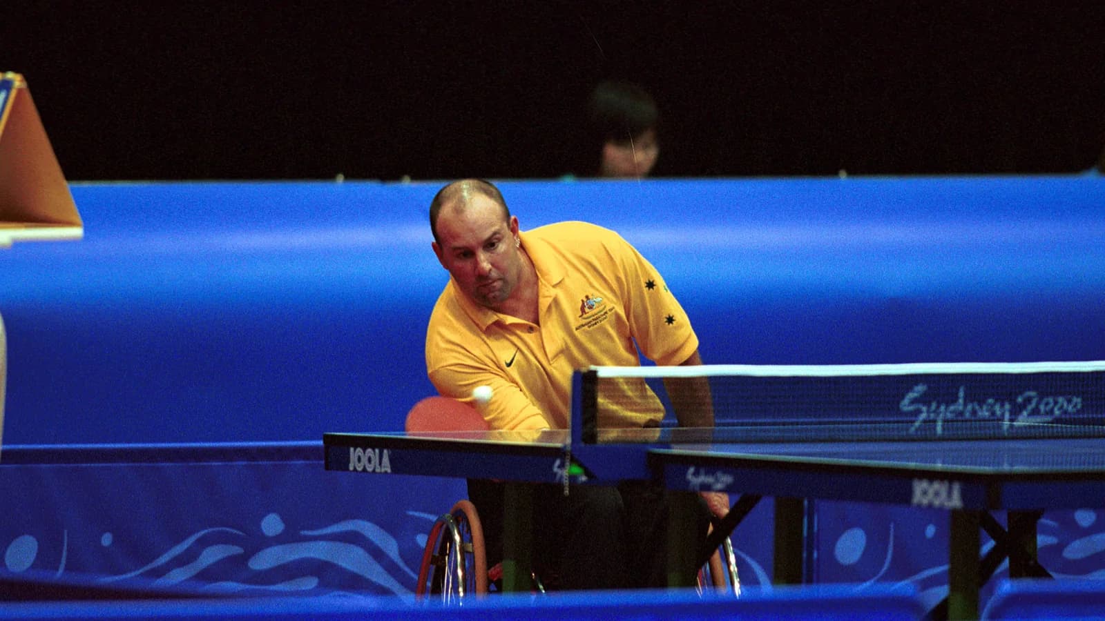 A table tennis player in a wheelchair serving at an indoor competition table, an opponent behind, under stadium lighting.