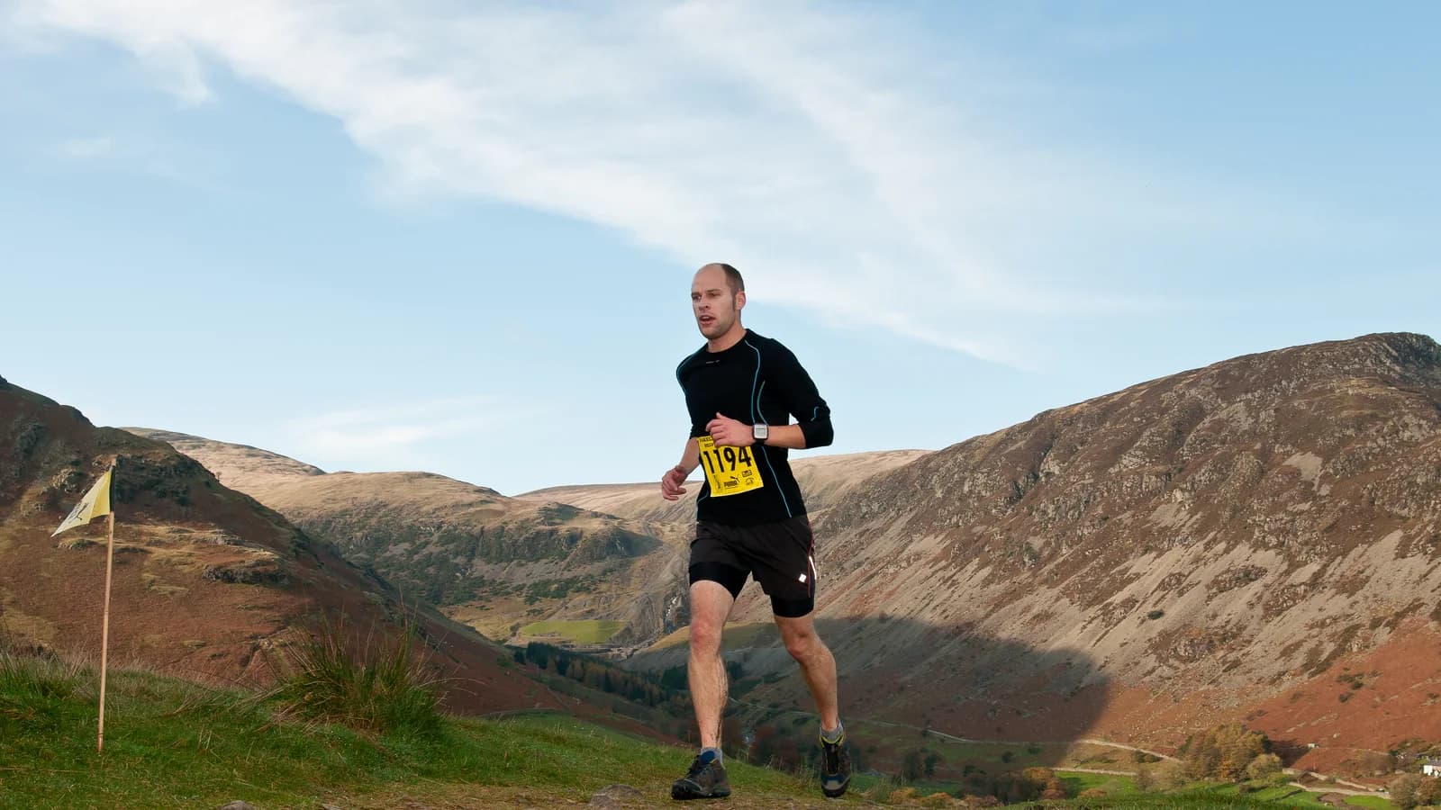 A runner wearing a race number crosses a grassy hillside path, with a broad valley and a steep fellside rising behind.