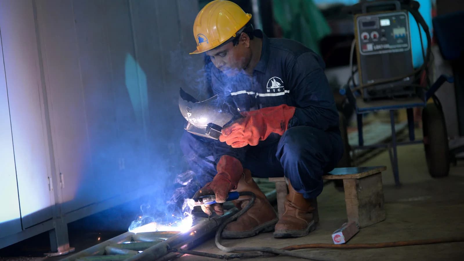 A welder in a hard hat, visor and gauntlets crouches over a bright arc, with smoke rising and a welding set behind.
