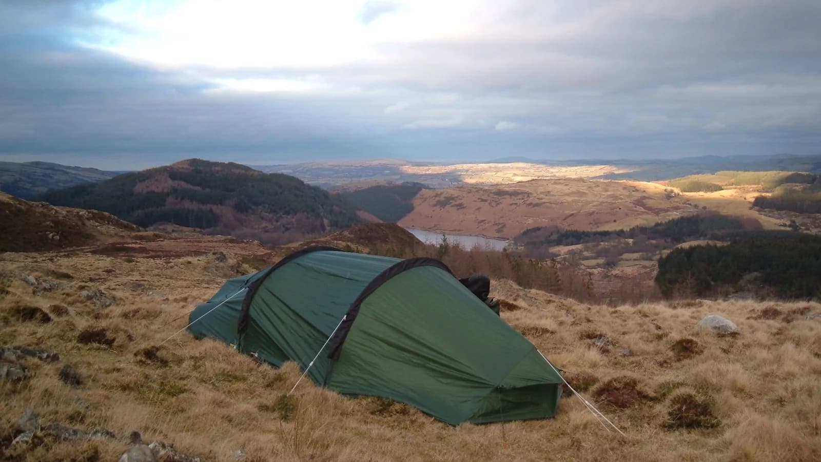 A small green tent pitched on rough moorland grass at golden hour, with a broad valley and hills falling away behind it.