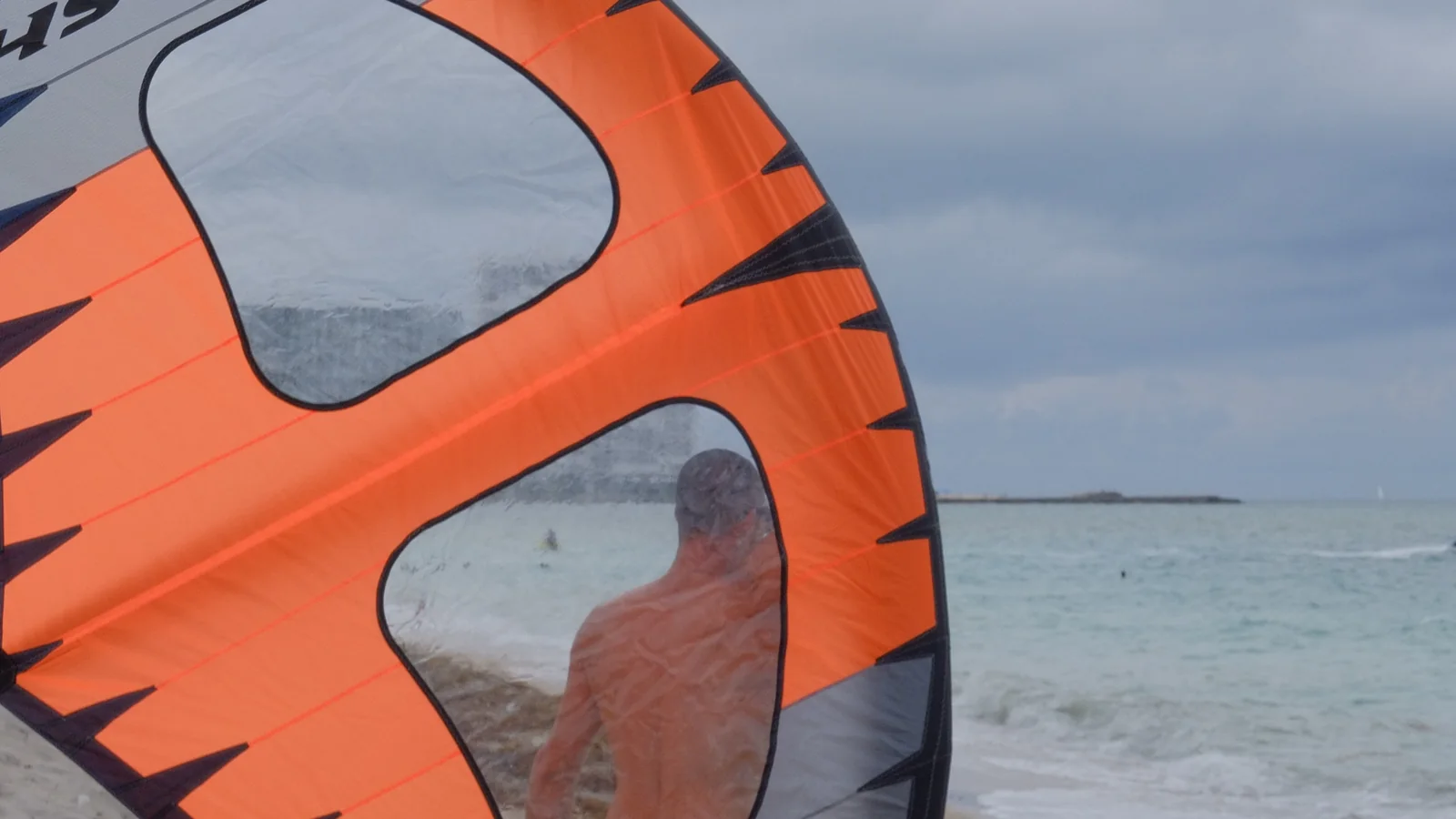 A rider standing in shallow water holding a large orange inflatable wing above the surface, seen through its window.
