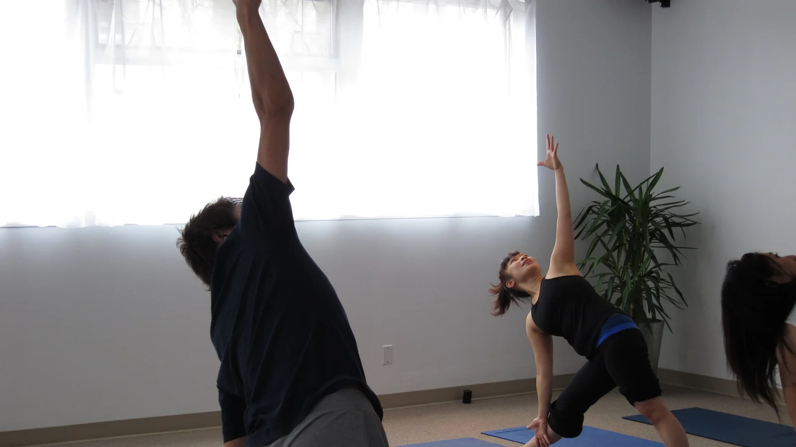 Three people on mats in a plain studio holding a standing side bend, one arm reaching overhead and the other to the floor, with daylight through a curtained window behind them.